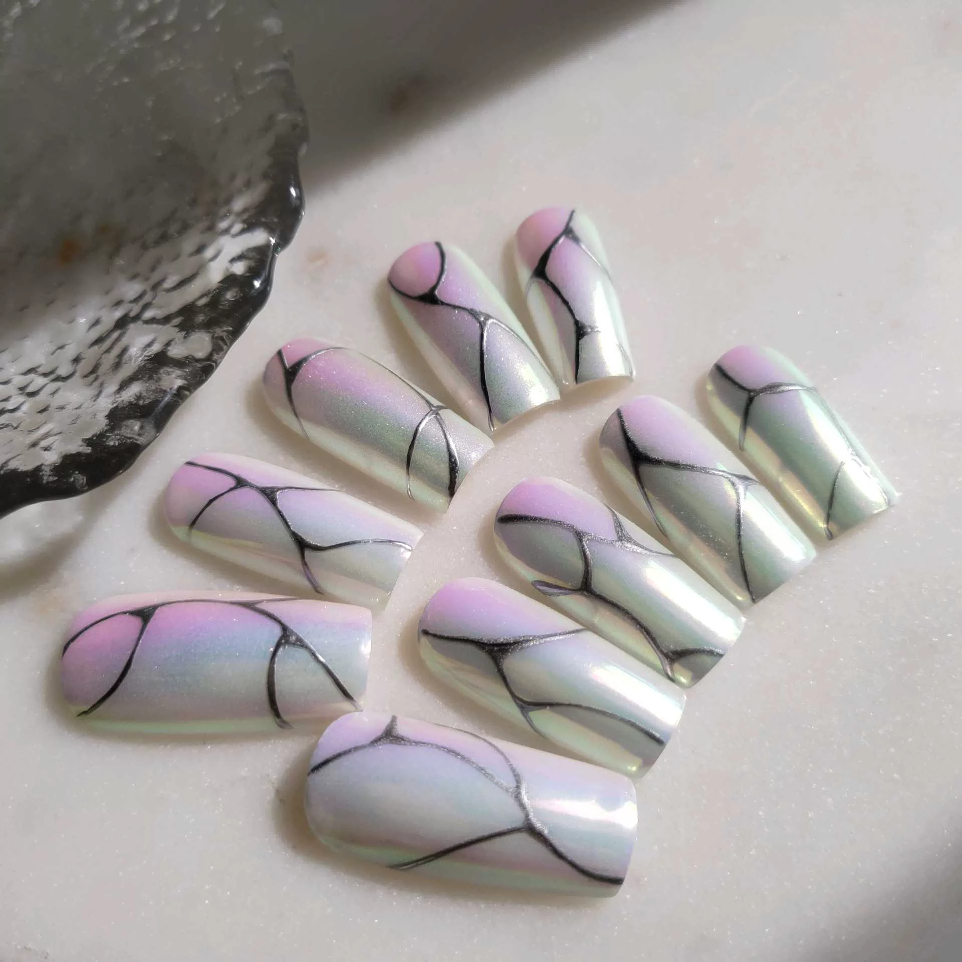A flat-lay display of medium square press-on nails featuring a shimmering, iridescent pearl chrome finish over a soft pink and mint green pastel gradient base, detailed with intricate, organic fine-line black abstract cracks or marble patterns.
