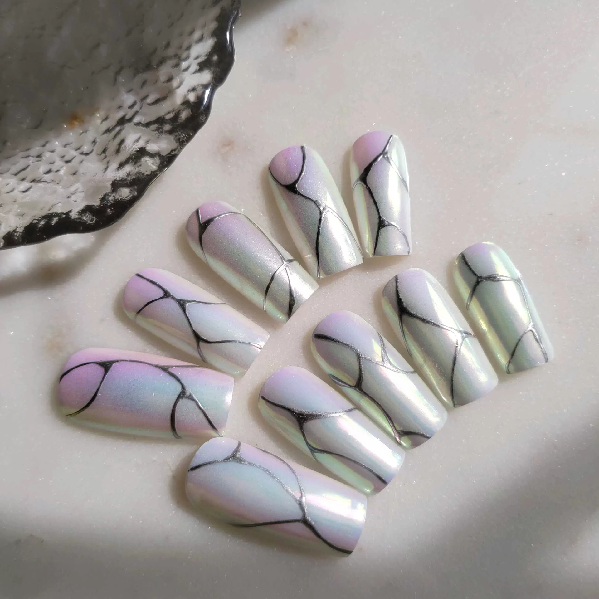 A flat-lay display of medium square press-on nails featuring a shimmering, iridescent pearl chrome finish over a soft pink and mint green pastel gradient base, detailed with intricate, organic fine-line black abstract cracks or marble patterns. — photo 2