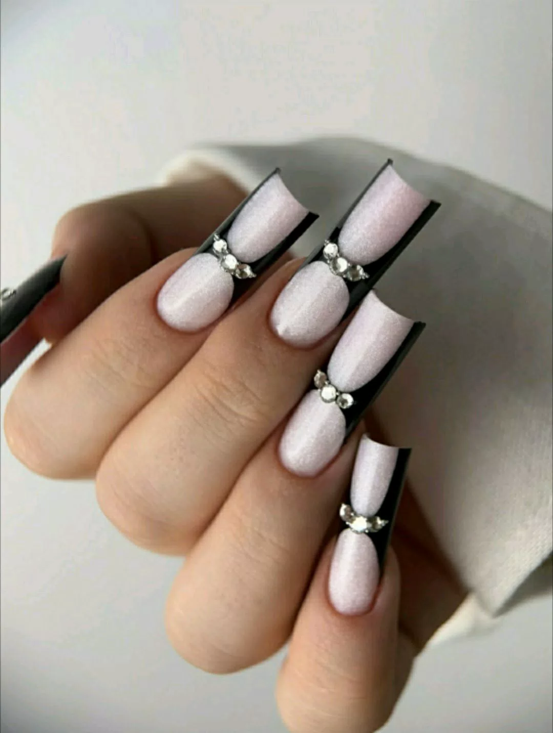 A close-up of a hand showcasing extra-long, square press-on nails featuring a shimmering pearlescent white base, deep black V-cut French tips, and a row of sparkling rhinestones accenting the smile line of each nail. — photo 2
