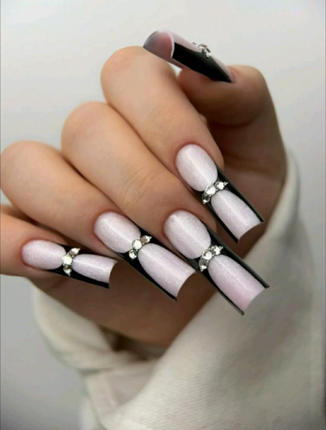 A close-up of a hand showcasing extra-long, square press-on nails featuring a shimmering pearlescent white base, deep black V-cut French tips, and a row of sparkling rhinestones accenting the smile line of each nail.