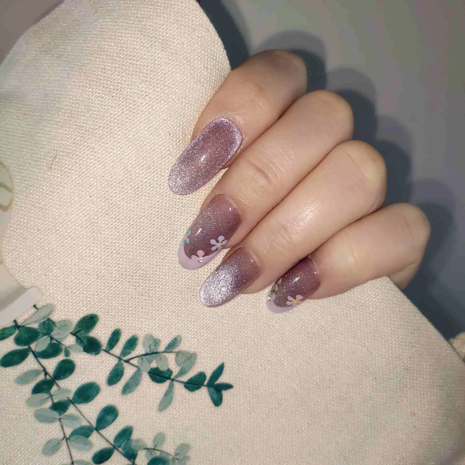A close-up of a hand showcasing long, oval press-on nails with a sheer, sparkling mauve-pink cat-eye base and elegant pastel pink French tips, delicately decorated with tiny hand-painted blue and white flowers and miniature rhinestones. — photo 3