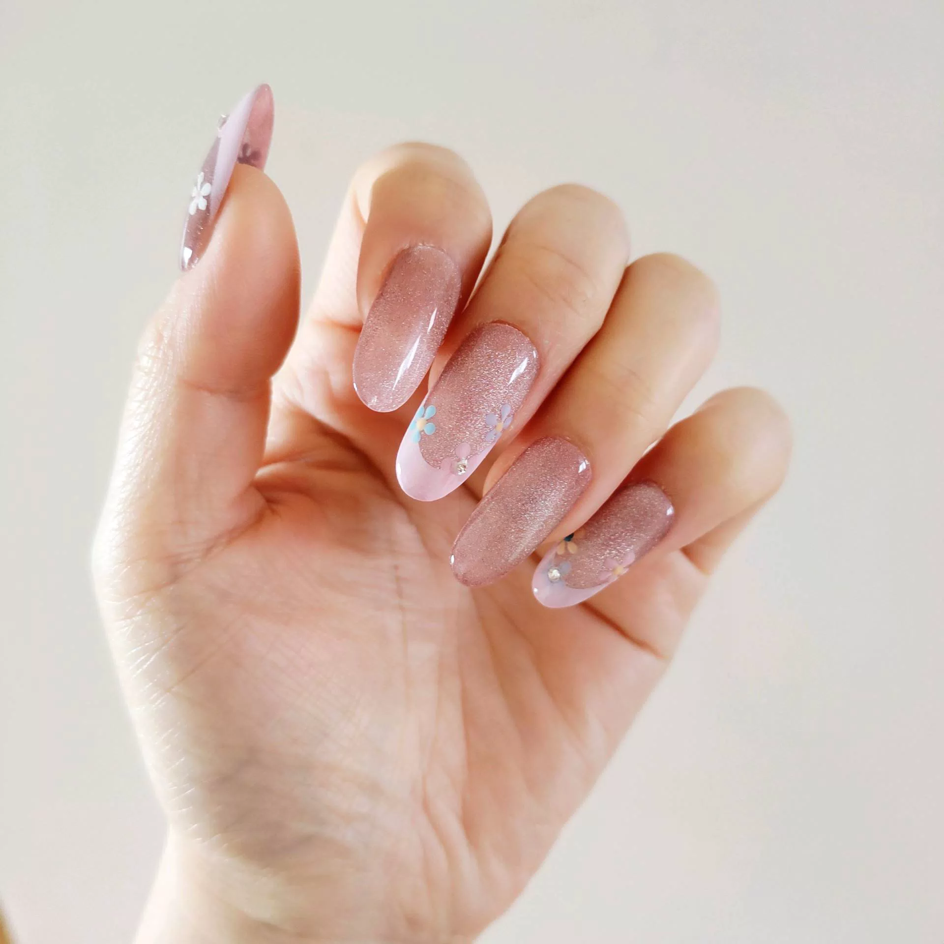 A close-up of a hand showcasing long, oval press-on nails with a sheer, sparkling mauve-pink cat-eye base and elegant pastel pink French tips, delicately decorated with tiny hand-painted blue and white flowers and miniature rhinestones.