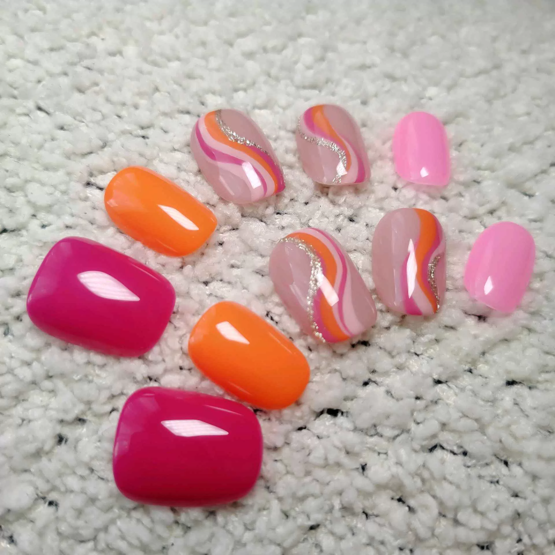 A hand showcasing short, rounded oval press-on nails with a mix of solid vibrant orange, hot pink, and abstract wavy swirl patterns in pink, orange, and white over a natural nude base. — photo 3