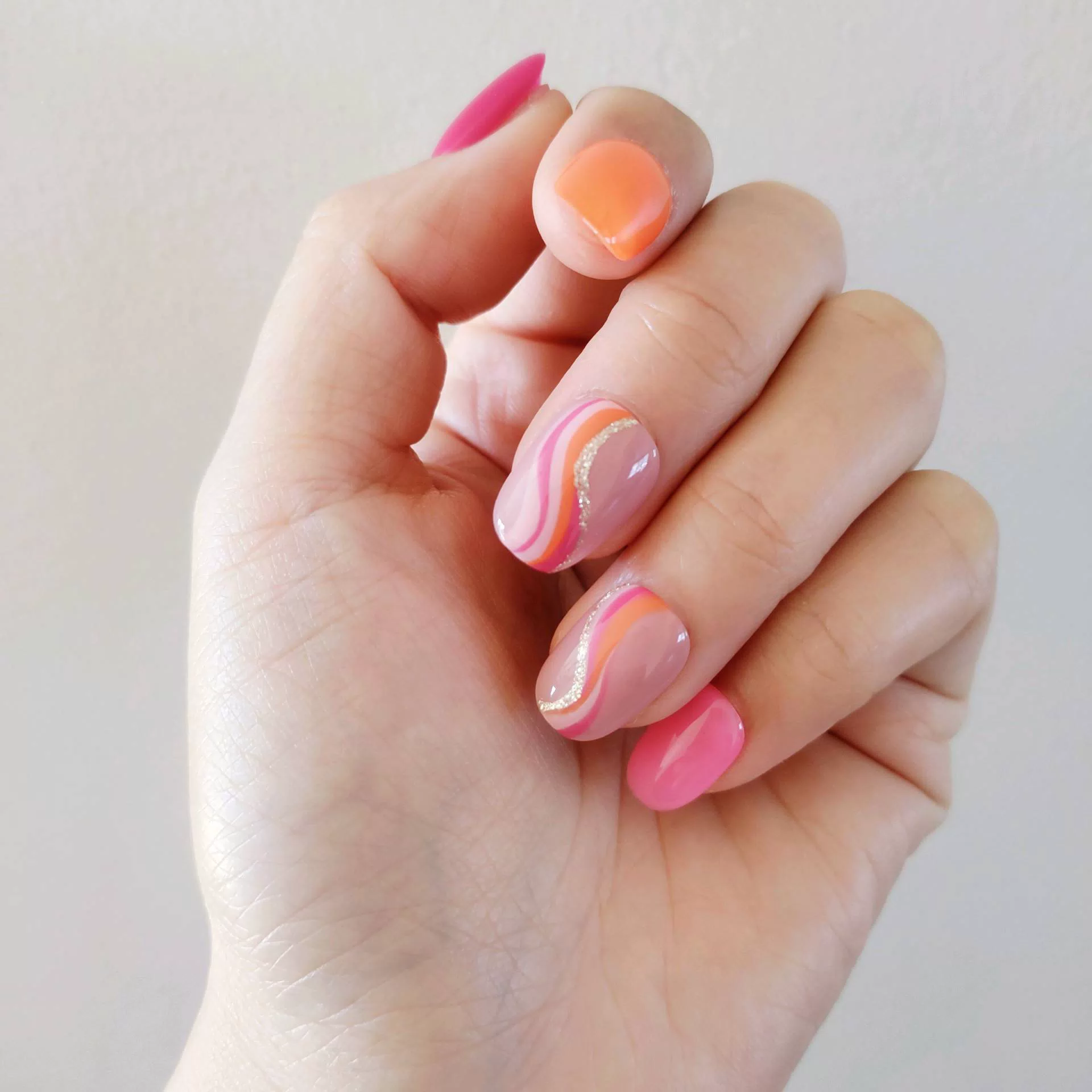 A hand showcasing short, rounded oval press-on nails with a mix of solid vibrant orange, hot pink, and abstract wavy swirl patterns in pink, orange, and white over a natural nude base.