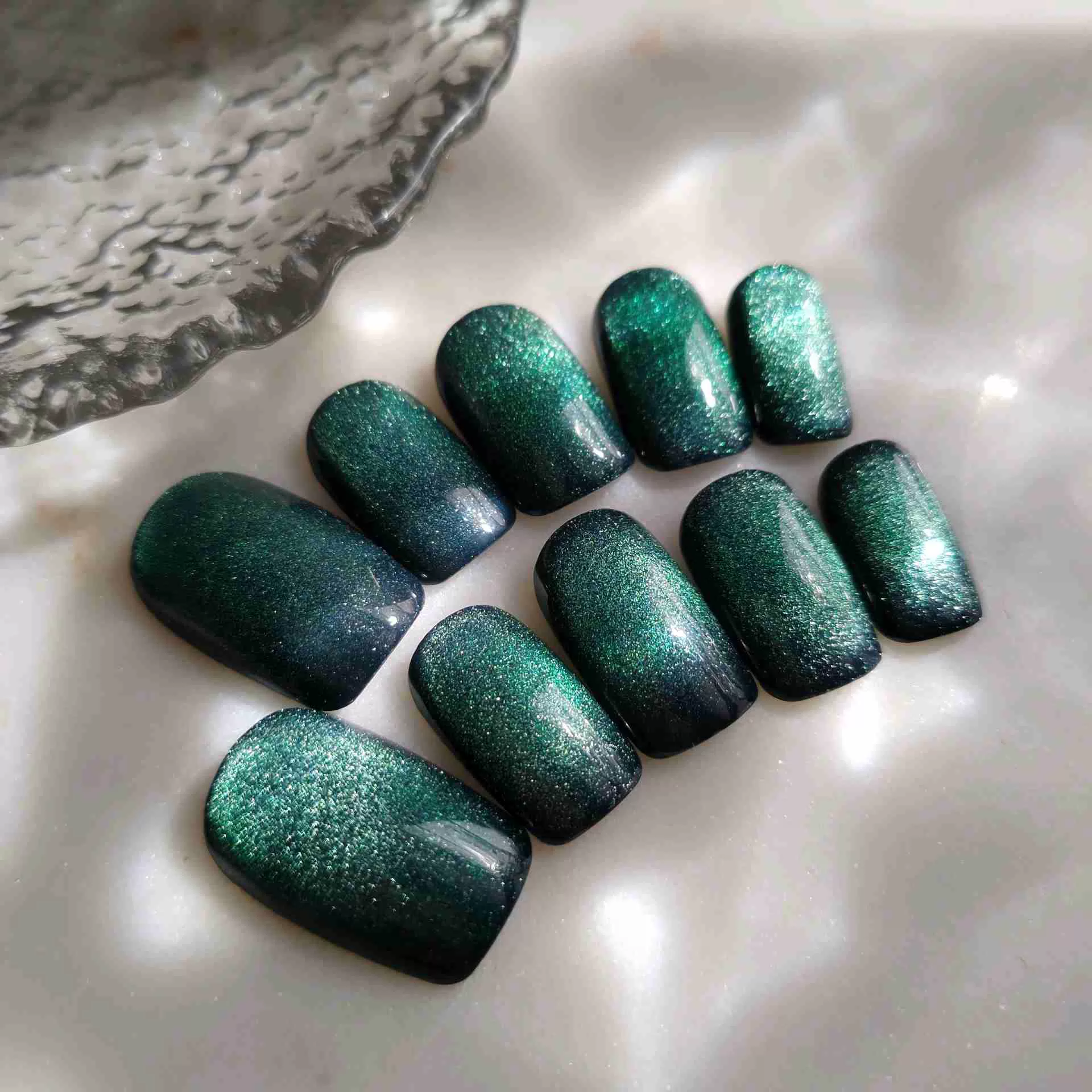 A professional top-down photograph displays a set of ten press-on nails arranged on a light grey, polished marble background. The nails are a short, functional square shape. They feature a mesmerizing deep forest green gel base activated with magnetic cat-eye pigment, creating a shimmering, velvet texture that shifts between dark emerald and vibrant teal. Soft lighting emphasizes the reflective, high-gloss finish of the nails.