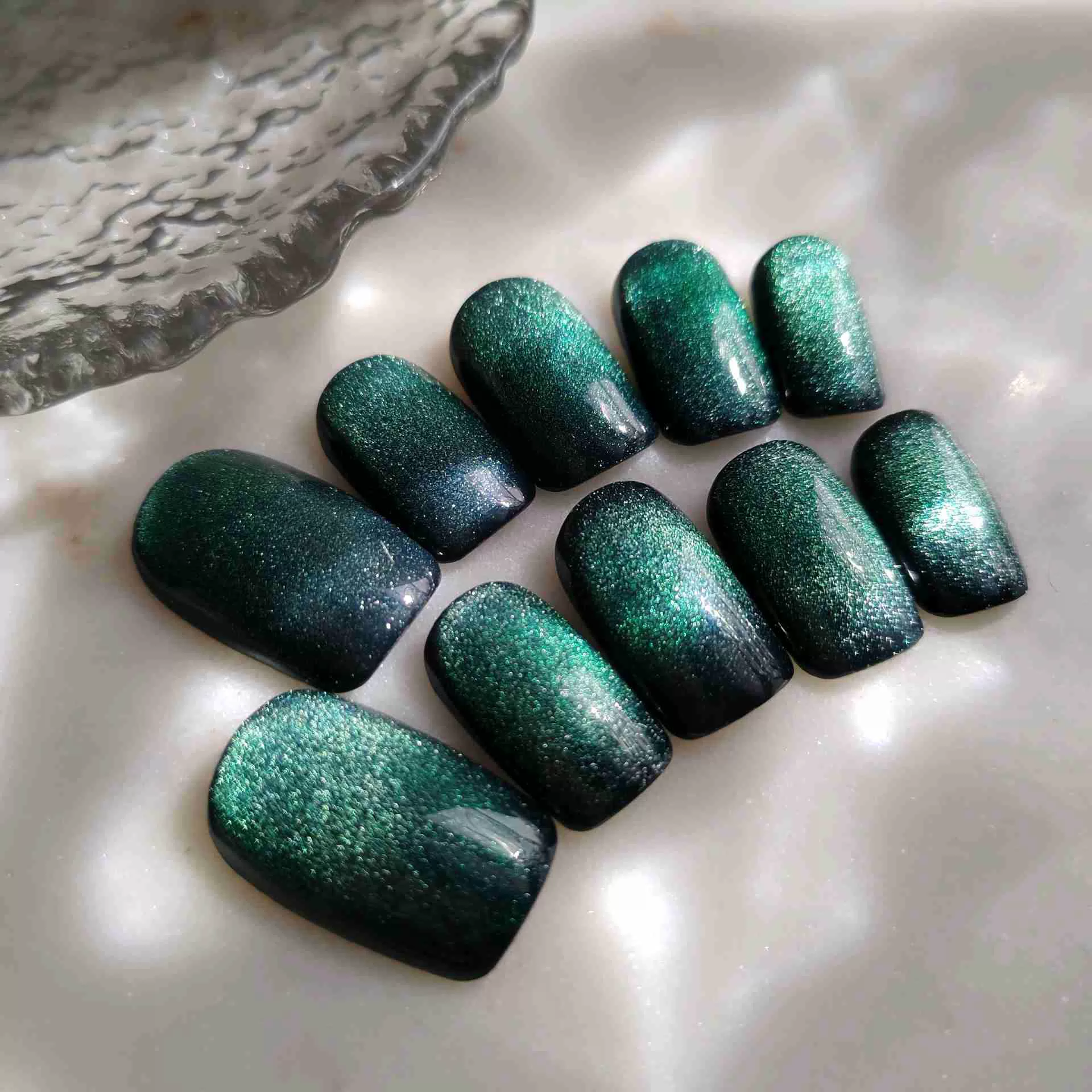 A professional top-down photograph displays a set of ten press-on nails arranged on a light grey, polished marble background. The nails are a short, functional square shape. They feature a mesmerizing deep forest green gel base activated with magnetic cat-eye pigment, creating a shimmering, velvet texture that shifts between dark emerald and vibrant teal. Soft lighting emphasizes the reflective, high-gloss finish of the nails. — photo 2