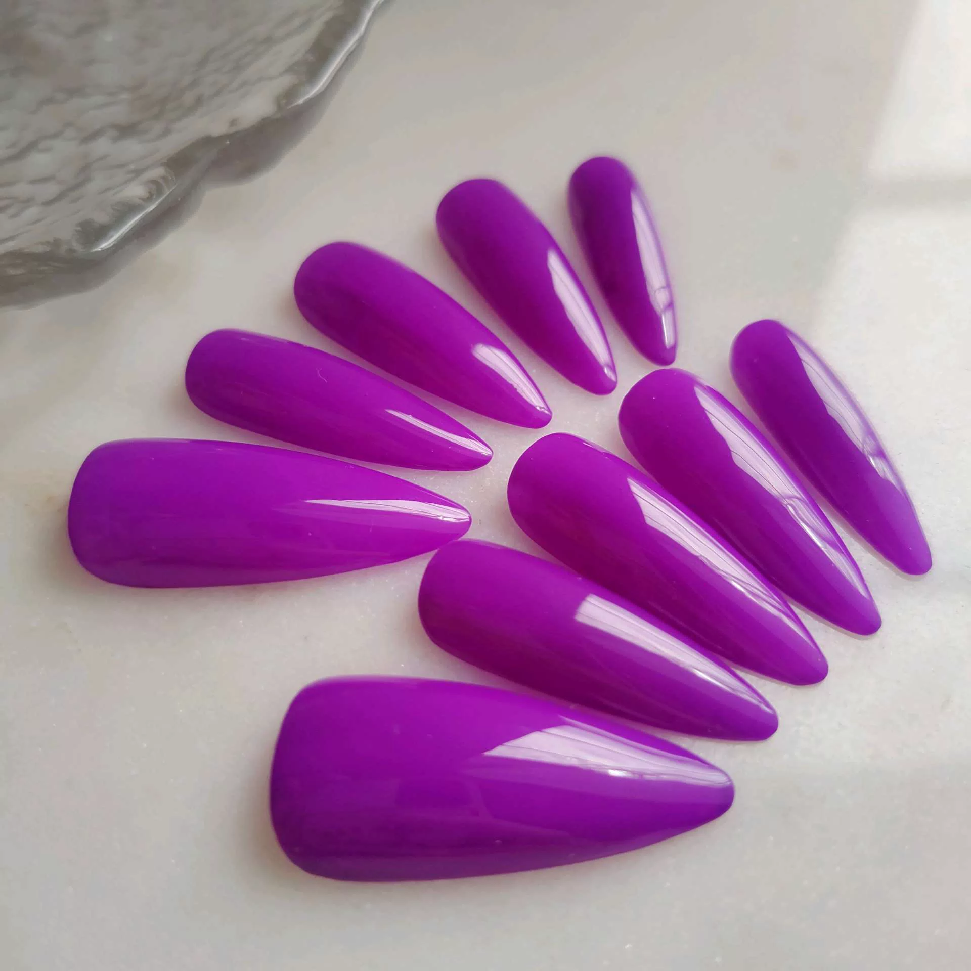 A flat-lay display of long, tapered almond-stiletto press-on nails in a solid, vibrant, ultra-glossy magenta purple color.