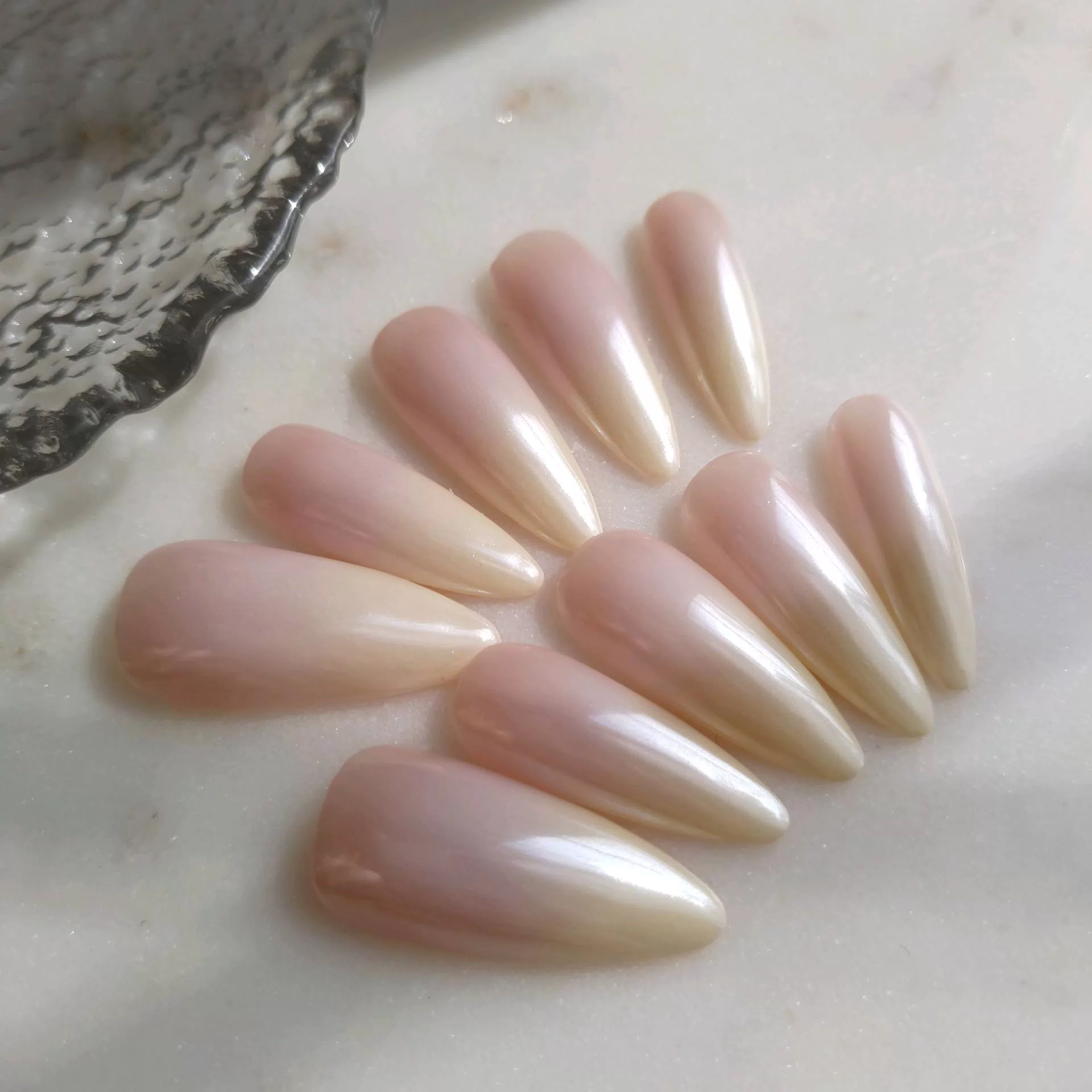 A flat-lay display of long, tapered almond press-on nails featuring a soft vanilla-nude ombre base coated in a high-shine, iridescent pearl chrome glaze.
