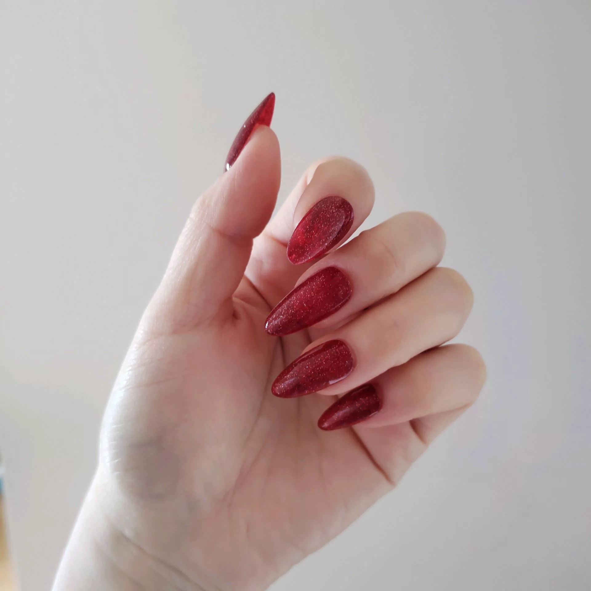 A close-up of a hand showcasing long, tapered stiletto-almond press-on nails coated in a deep, luxurious ruby red magnetic cat-eye shimmer that reflects light with a rich, multi-dimensional velvet finish.