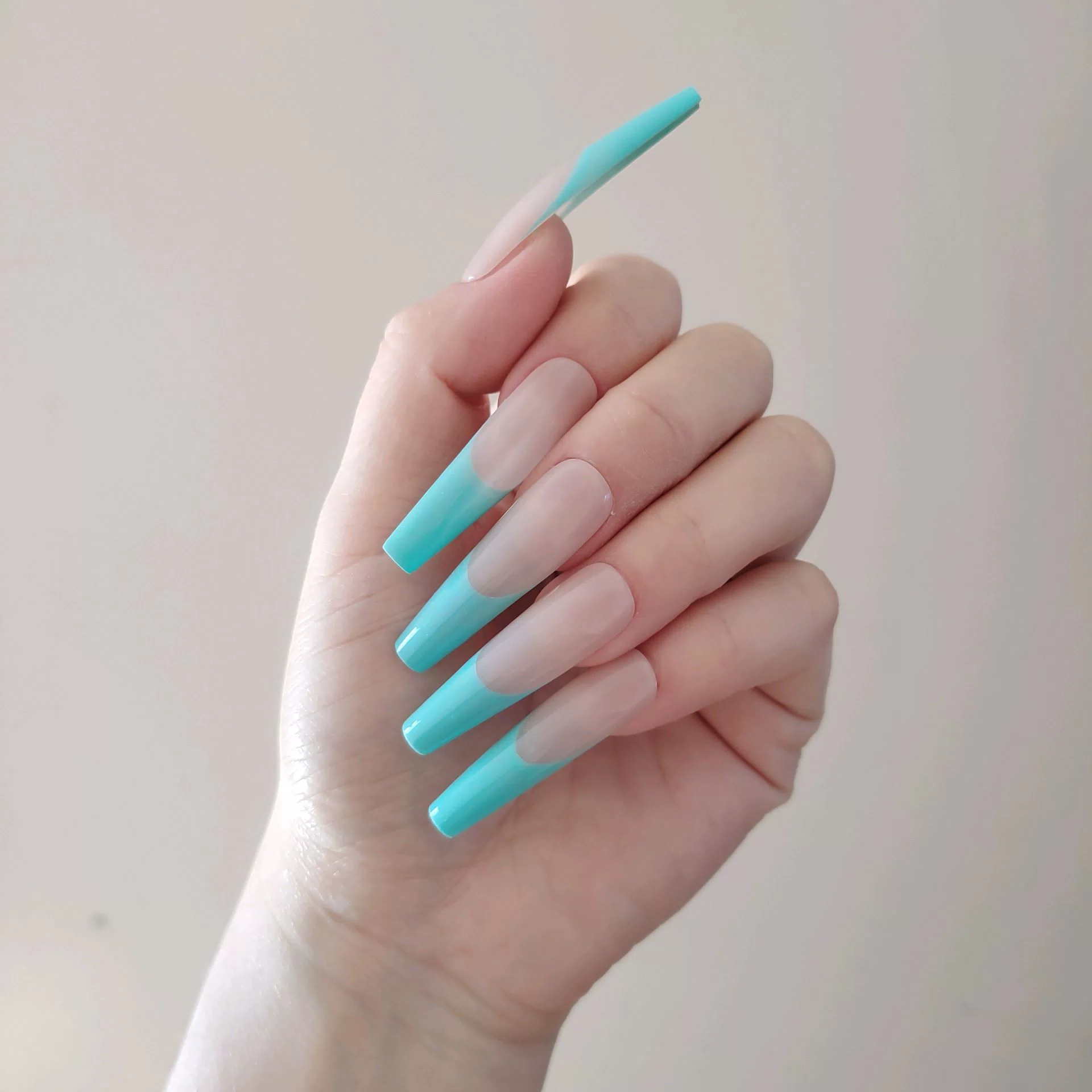 A close-up of a hand showcasing extra-long, square press-on nails featuring a matte nude base and vibrant turquoise-teal French tips aligned in a clean, high-contrast smile line.