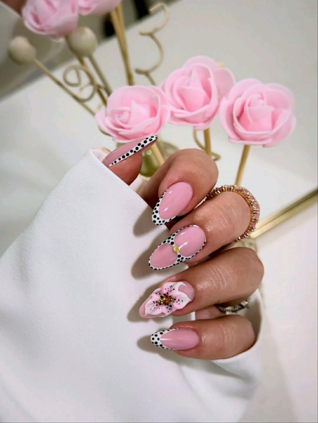 A close-up of a hand showcasing medium almond-shaped press-on nails with a glossy pink base. The tips feature a black and white dalmatian animal print French style, with a large, handcrafted 3D pink and white flower on the ring finger accented by a sparkling amber rhinestone center.