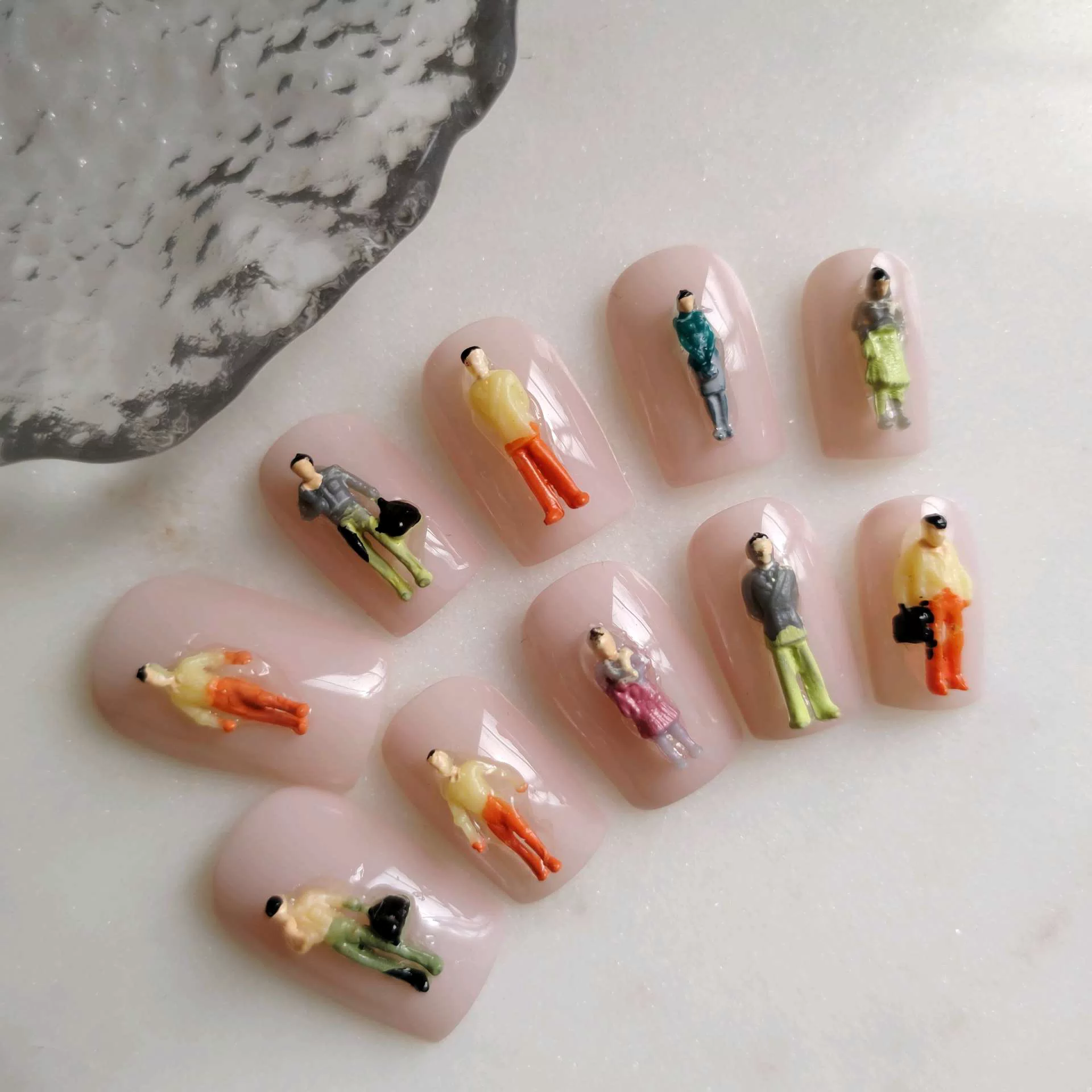 A flat-lay display of short, square press-on nails with a smooth, glossy nude-pink base, each uniquely adorned with a highly detailed, miniature 3D architectural model figure of a person standing or walking in colorful clothing. — photo 2
