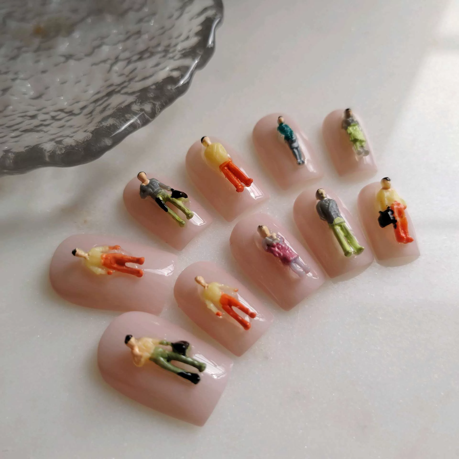A flat-lay display of short, square press-on nails with a smooth, glossy nude-pink base, each uniquely adorned with a highly detailed, miniature 3D architectural model figure of a person standing or walking in colorful clothing.