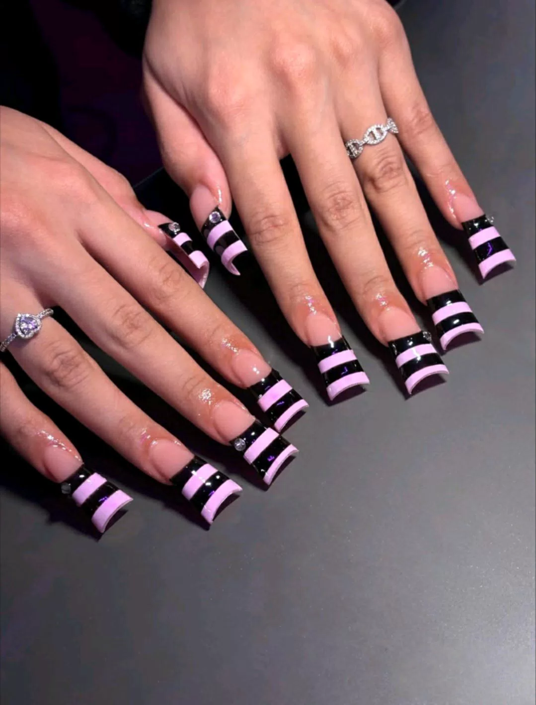 A close-up of two hands showing extra-long, wide flared duck-style press-on nails with a nude base, a sharp black smile line, and alternating horizontal pink and black stripes on the tips, accented by tiny silver rhinestones. — photo 2